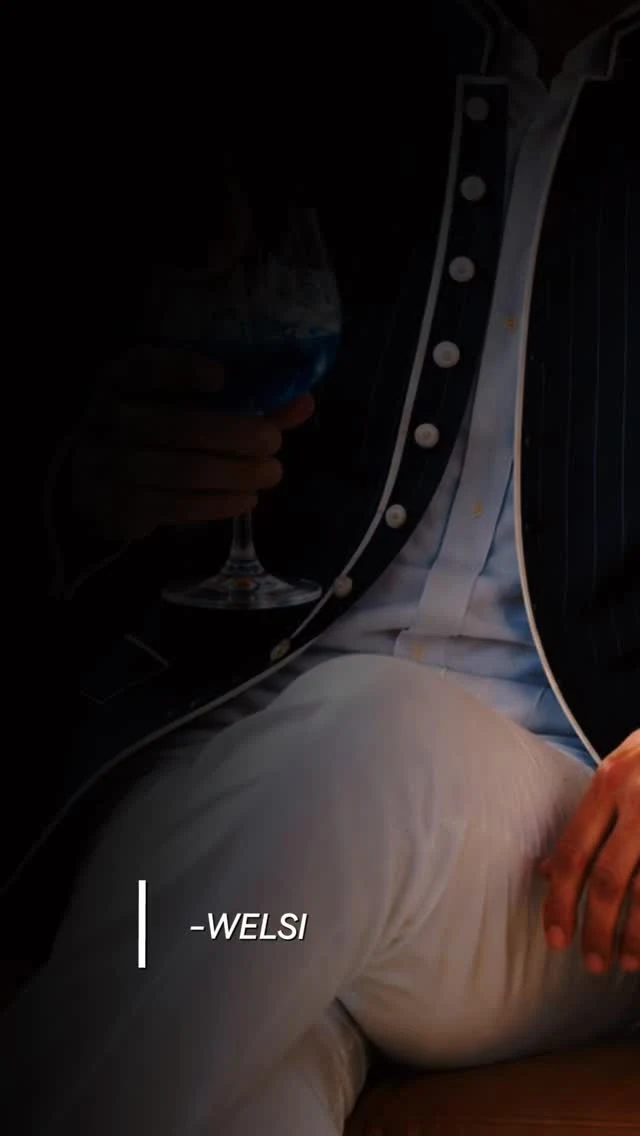 Americana oscura de raya diplomática y pantalón blanco, con una copa azul en la mano