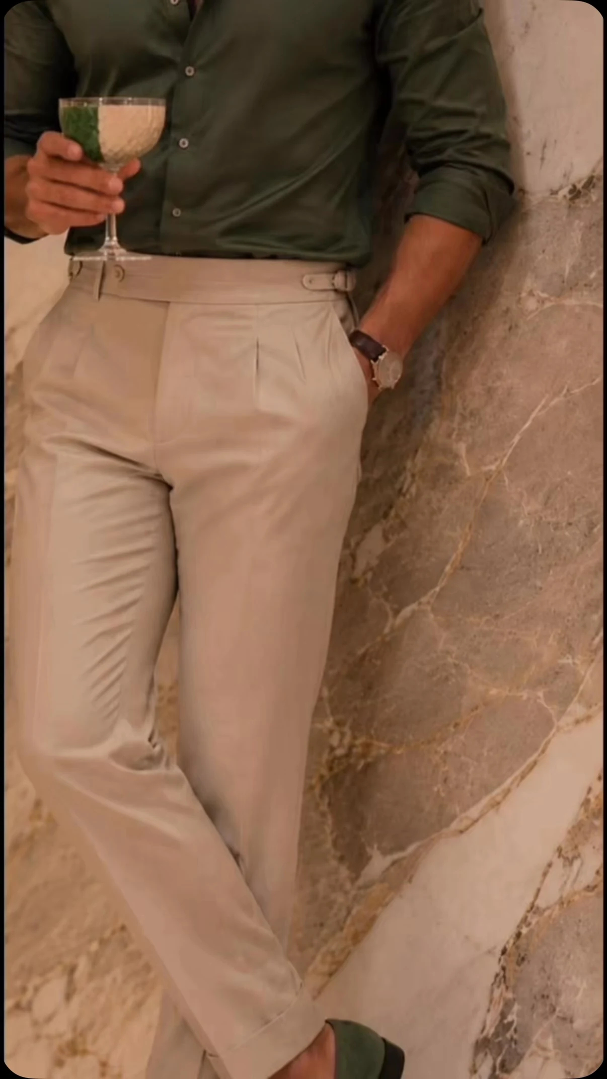 Camisa verde y pantalón beige de pinzas, con un cóctel en la mano junto a una pared de mármol